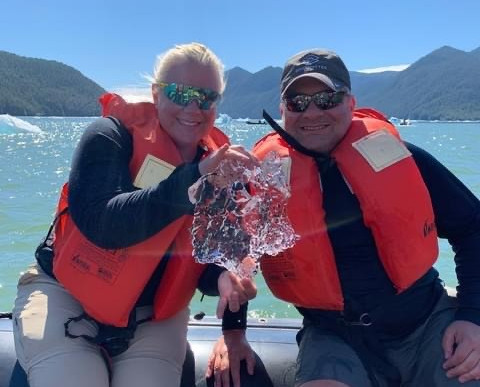 A woman and man in orange life jackets sit on a boat, smiling and holding up a piece of glacial ice between them, with icebergs, water, and forested mountains in the background.