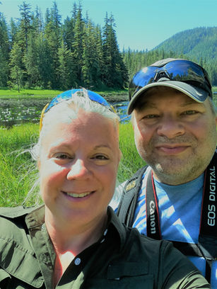 Two people smiling for a selfie in front of a peaceful forest lake surrounded by evergreen trees and rolling hills on a sunny day.