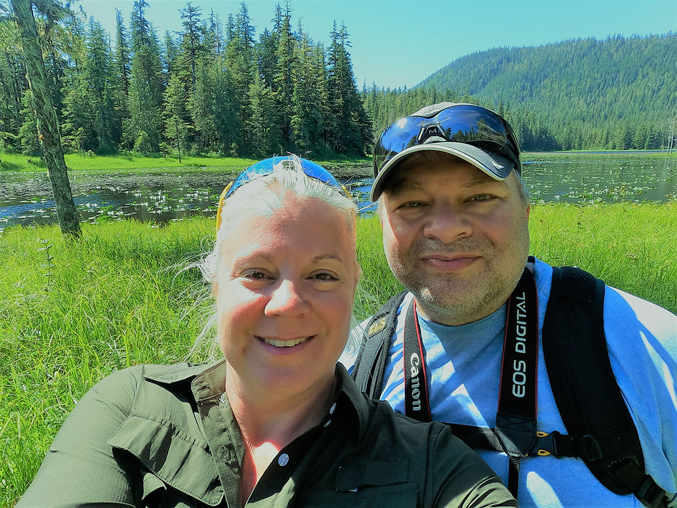 Two people smiling for a selfie in front of a peaceful forest lake surrounded by evergreen trees and rolling hills on a sunny day.