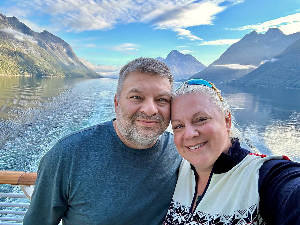 A man and woman lean their heads together, smiling for a selfie on the deck of a ship, with a scenic fjord, steep green-and-rocky mountains, and a distant snow-dusted peak behind them under a partly cloudy blue sky.
