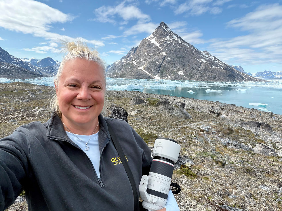 A woman wearing a "Glacial Expeditions" fleece takes a selfie while holding a large white camera lens, with a rocky landscape, icebergs floating in turquoise water, and a sharp mountain peak behind her.