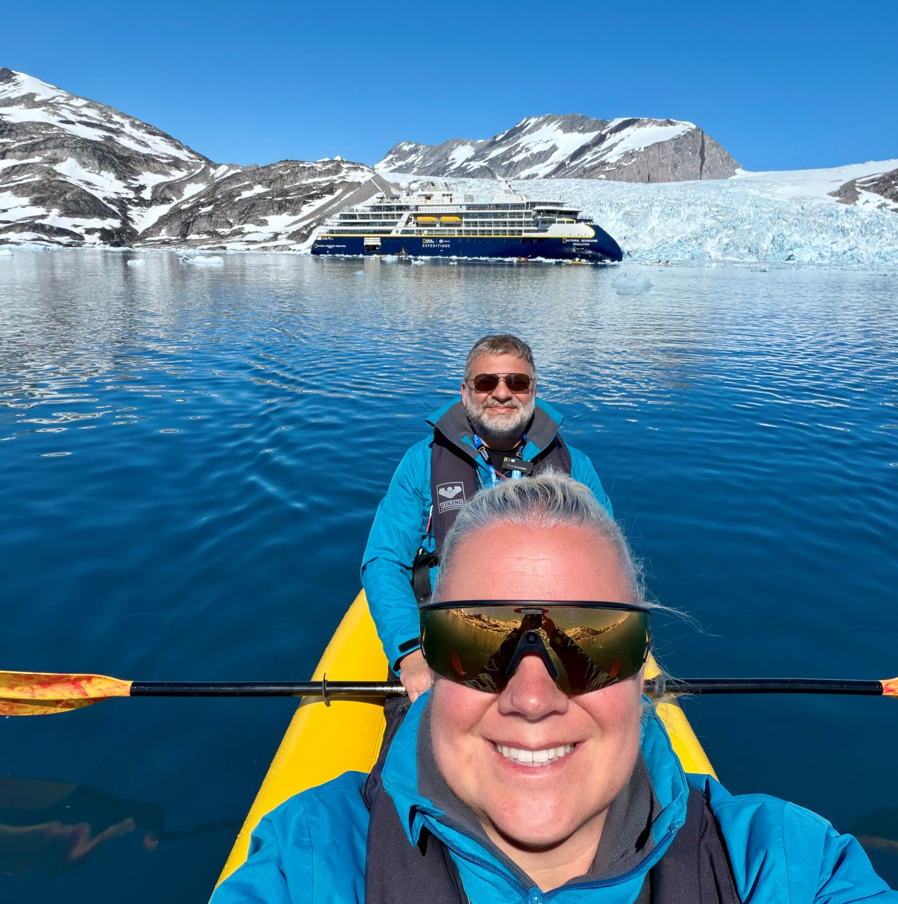 A woman and man in blue kayaking jackets sit in a yellow kayak on calm, deep-blue water, smiling for a selfie, with a large expedition ship, glacier, and snow-capped mountains in the background.