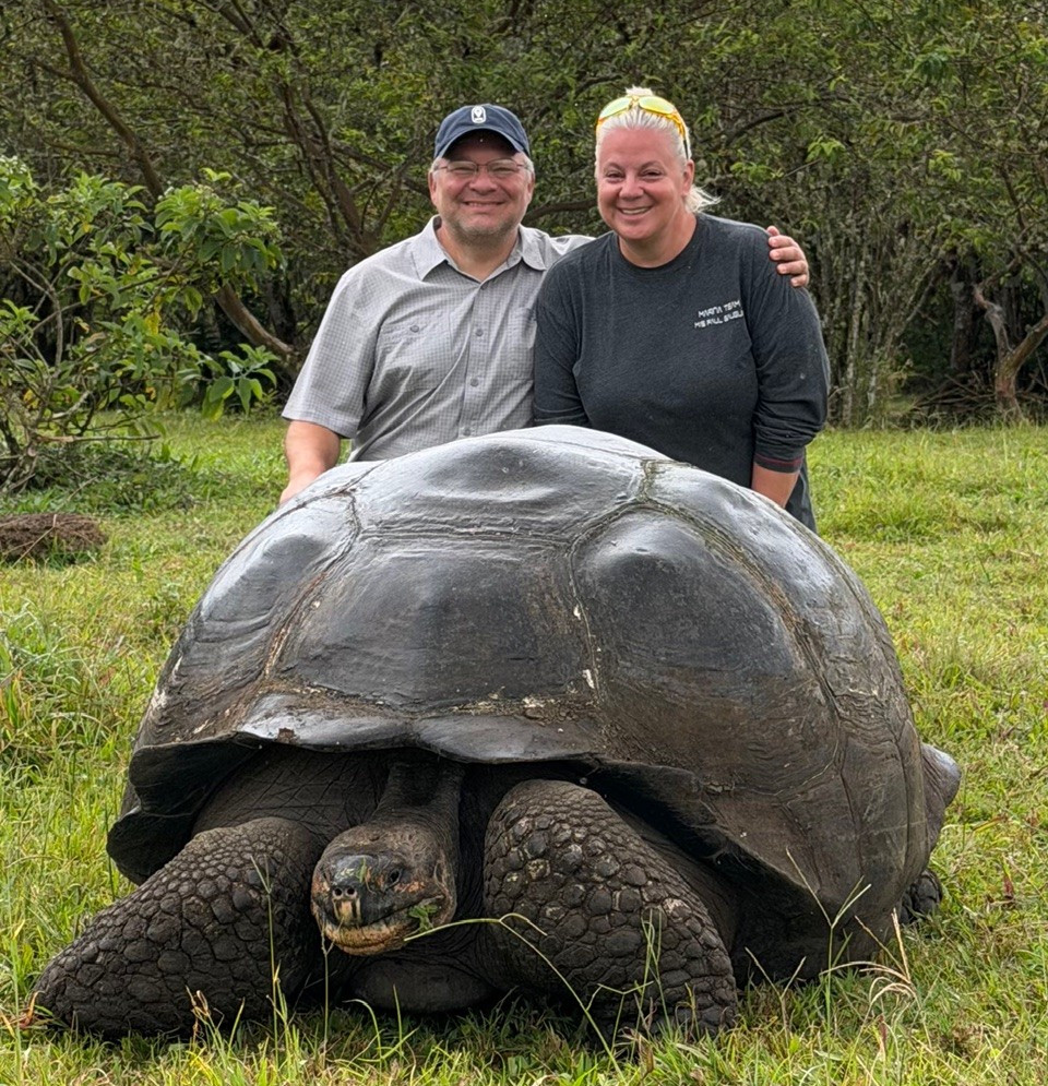 A woman and man stand smiling behind a large Galápagos giant tortoise on a grassy field, with dense green trees in the background.