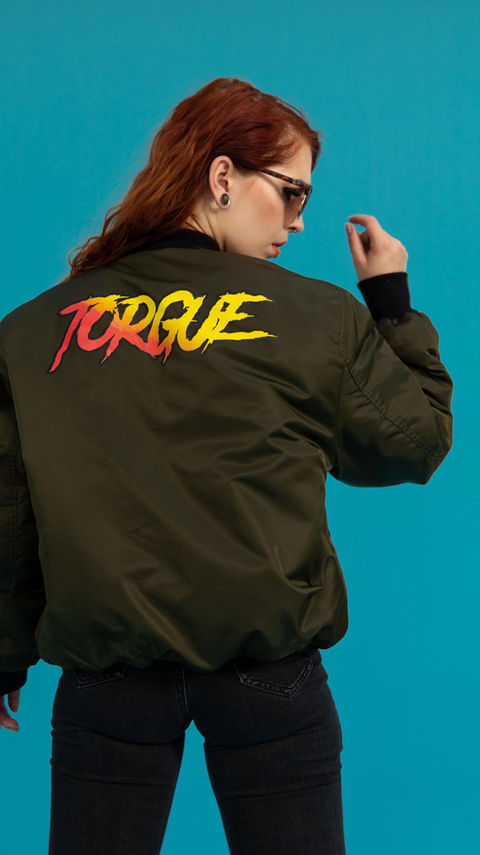 Carmen Hellena with a "Torque" jacket and red hair for a Borderlands 3 fashion shoot: Carmen Hellena, with her back to the camera, wears a black jacket with the word "Torque" in bold red letters, against a blue background. This edgy and cool fashion shoot is inspired by Borderlands 3.