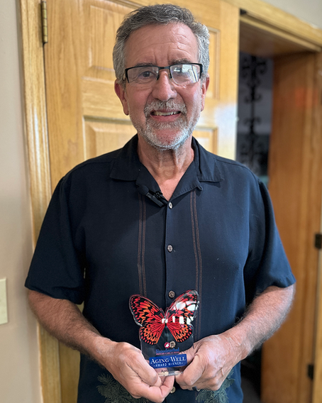 Kevin Talbot smiles while holding his Freudenthal Aging Well Award, standing in front of a wooden door, wearing a navy shirt with a butterfly-themed trophy in hand.