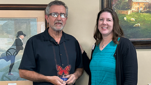 Kevin Talbot stands beside Leah from Freudenthal, holding his butterfly-shaped Aging Well Award, both smiling in front of framed artwork.
