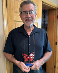 Kevin Talbot, smiling, stands with his Aging Well Award butterfly trophy, framed by warm indoor lighting and a wooden door in the background.