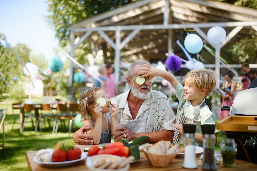 Grandfather laughing with two grandchildren at an outdoor summer picnic, surrounded by food and decorations.