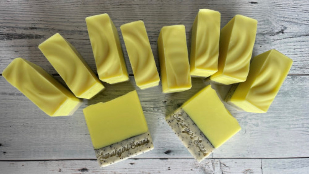 Top-down view of 10 square yellow bars of soap on pale wooden surface, 8 bars are standing on end showing ridges in surface, 2 bars lie flat showing bottom third is a tan color with black speckles and stamped with "Sarahgirl Soaps"
