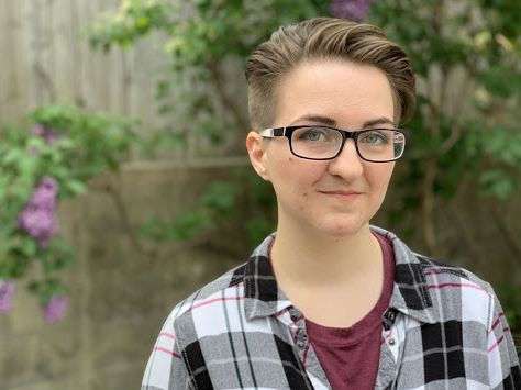 Headshot of Joehey Cici. Black, white, and maroon plaid collared shirt over maroon T-shirt. Black and white rectangular framed glasses. Wooden wall and greenery in background.