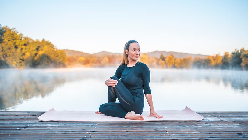 Woman in black yoga clothes sits on a mat on a wooden dock by a misty lake, with autumn trees and hills at sunrise.