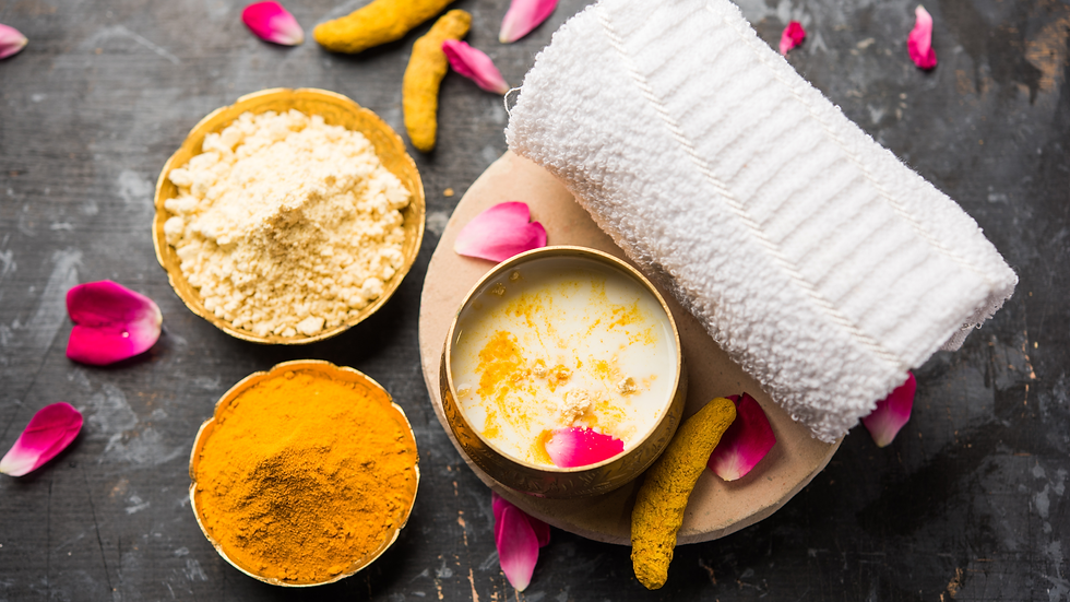 Spa setup with bowls of yellow and white powders, a cream in a brass cup, rolled white towel, and pink rose petals on dark wood.
