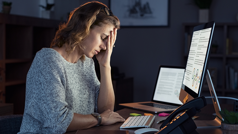A woman in a gray sweater looks stressed at her desk with a hand on her forehead. Two computer screens display documents. Dimly lit room.