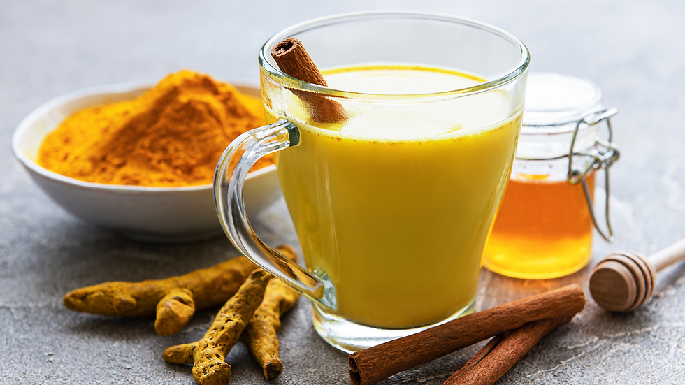 Glass of yellow turmeric milk with a cinnamon stick, bowl of turmeric powder, honey jar, cinnamon sticks, and turmeric roots on gray surface.