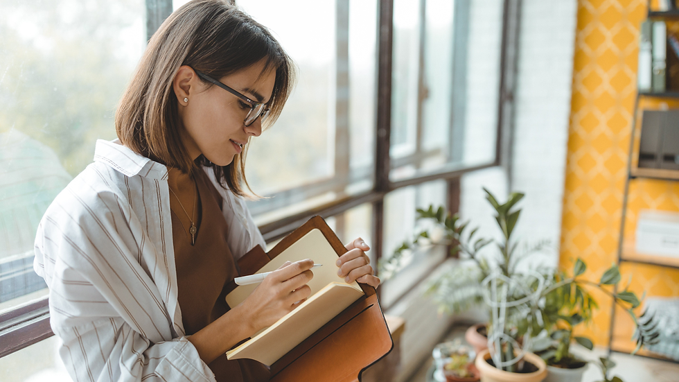 Woman with glasses writing in notebook by large windows, indoor plants, and yellow patterned wall. Bright, contemplative mood.