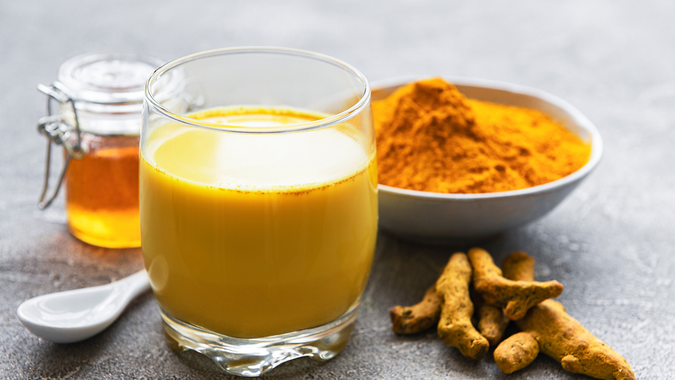 Turmeric latte in a glass with honey jar, spoon, turmeric powder bowl, and roots on a dark countertop.