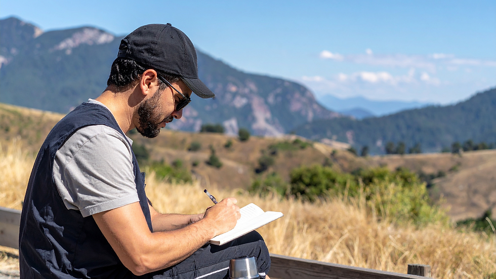 Man in black cap and sunglasses writes in a notebook on a sunny mountainside, with blue sky and rugged hills behind him.
