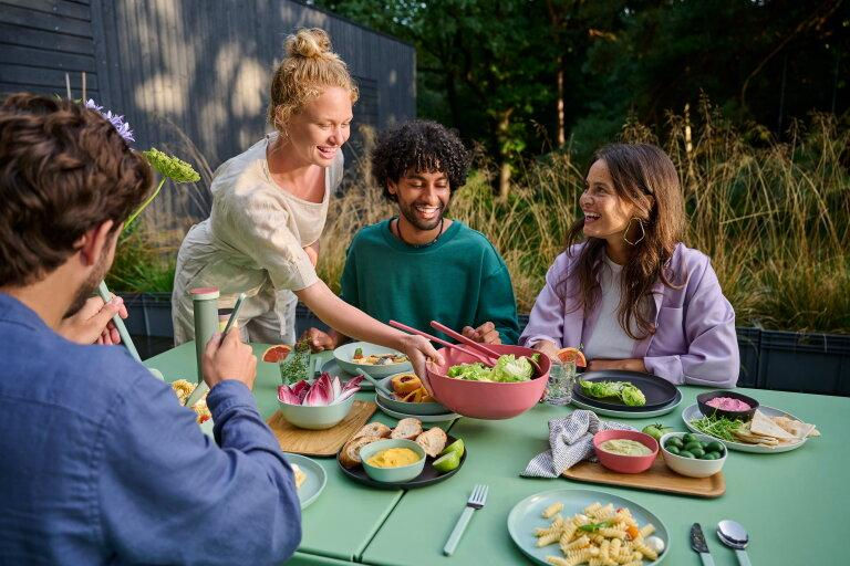 Friends laugh at an outdoor meal as a woman serves salad at a green table with colorful bowls.