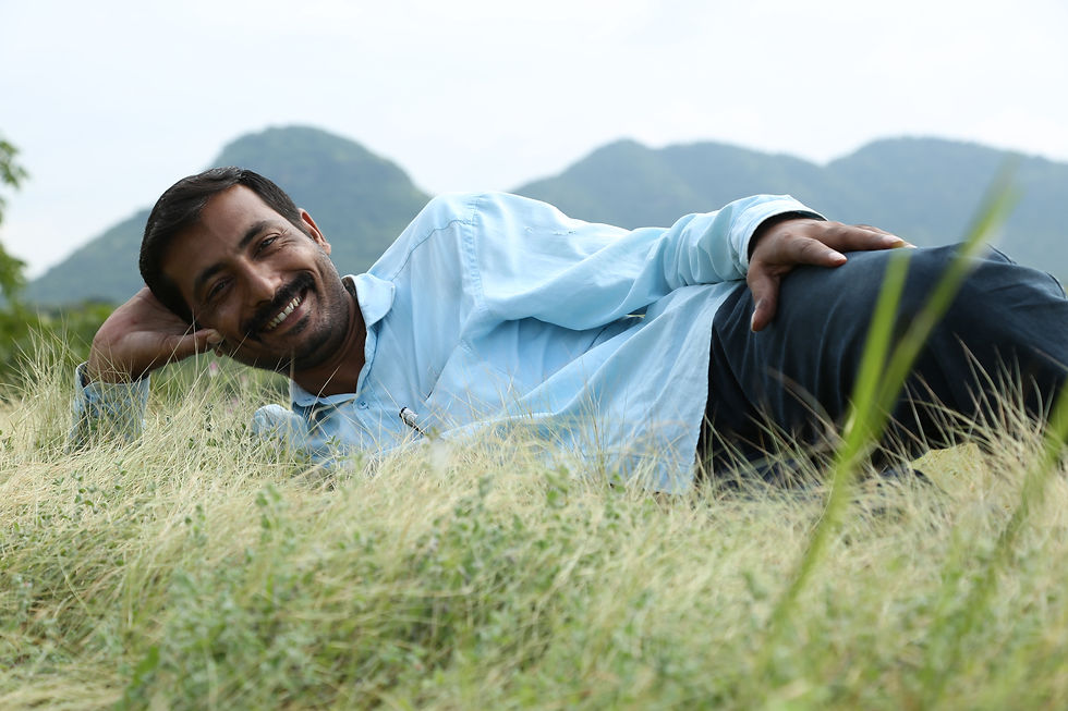 Man in a blue shirt smiles while lying on grass with hills in the background. The scene is calm and natural.