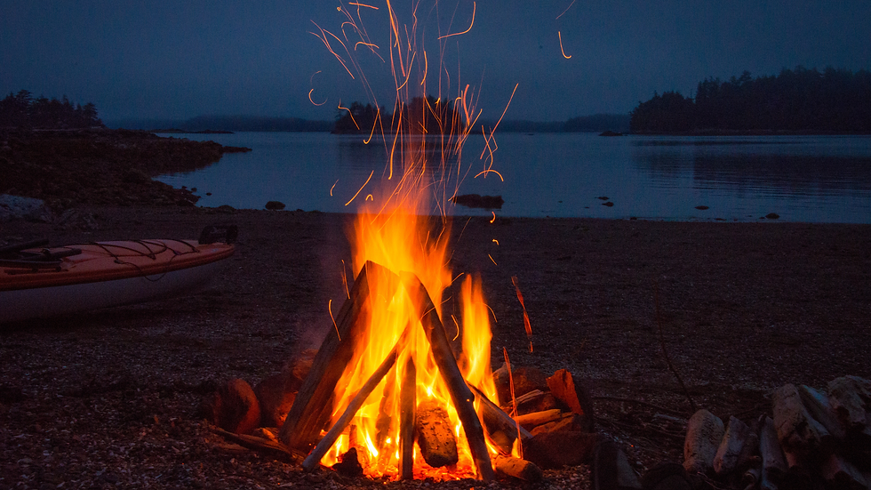 Campfire on a beach at dusk, flames and sparks rise against a dark, serene lake and tree silhouette, with a kayak nearby.