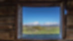 View of snow-covered mountains through a rustic wooden window. Green fields and blue sky create a serene, natural scene.