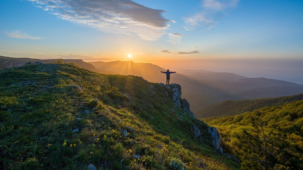 Person stands on a cliff with arms outstretched at sunrise over misty mountains and grassy hills.