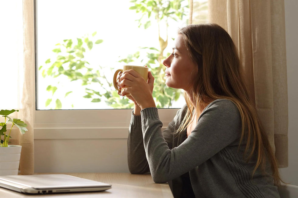 Woman in a gray sweater holds a mug by a sunlit window, gazing outside beside a laptop and potted plant.