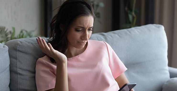 Woman in a pink shirt looks perplexed at her phone while sitting on a gray couch. She raises one hand in frustration. Indoor setting.