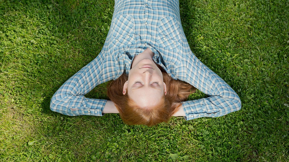 A person in a blue plaid shirt lies relaxed on green grass with eyes closed, arms behind head, creating a serene, peaceful mood.