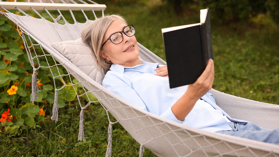 Woman in glasses reads a book while lying in a hammock outdoors. She's wearing a blue shirt, surrounded by greenery and orange flowers.
