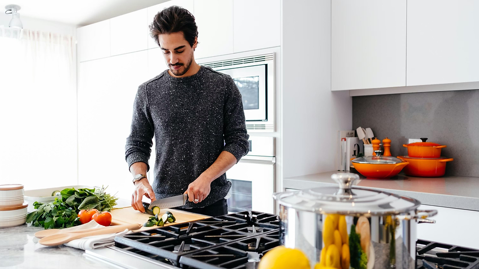 Man slices zucchini at a kitchen island in a bright white kitchen, with tomatoes, greens, and orange pots nearby.