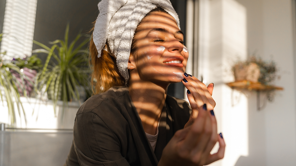 Woman with towel-wrapped hair applies face cream in warm sunlight, smiling in a bathroom with plant shadows.