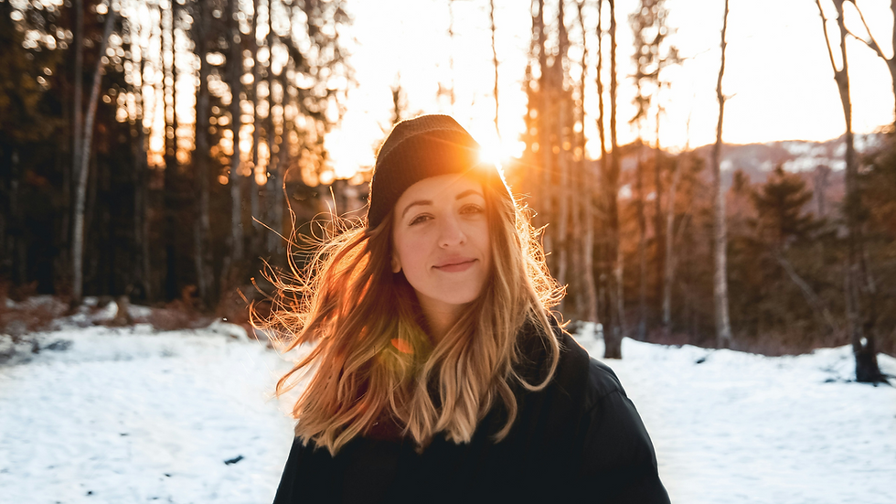Woman in a black beanie smiling in snowy forest at sunset. Sunlight glows through trees, creating a warm, serene atmosphere.