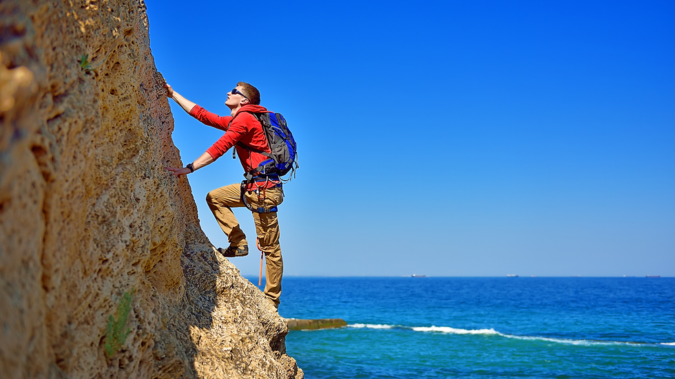 Climber in red hoodie and backpack scales a rocky seaside cliff above a blue ocean under a clear sky.