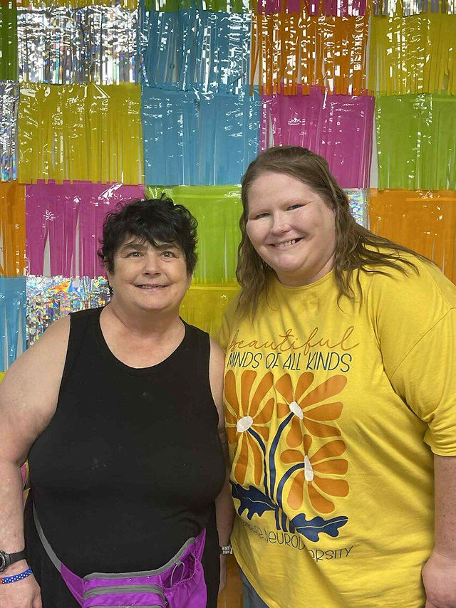 Two smiling individuals stand in front of a colorful, foil fringe backdrop. One wears a black tank top, the other a bright yellow shirt with flower graphics.