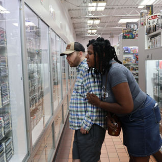 A man in a plaid shirt and cap, and a woman in a gray shirt and denim skirt, intently browse CDs in a music store. Shelves are filled with albums.