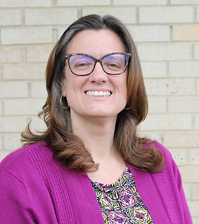 Smiling woman with glasses, long brown hair, wearing a purple sweater over a patterned blouse, stands against a beige brick wall.