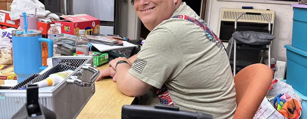 The image depicts a person seated on an orange office chair at a cluttered desk in a room with various items. The individual is wearing a beige cap and a light grey T-shirt, and is turned slightly towards the camera with a smile. The desk is crowded with a variety of objects such as a water bottle featuring a cartoon character, snack boxes, and small containers. Behind the person, a refrigerator with several photos and a list is positioned next to a partially open door leading to a dark corridor. A wall-mounted clock and a chalkboard are visible, displaying several pieces of chalk writing. Storage boxes and drawers can be seen in the background.