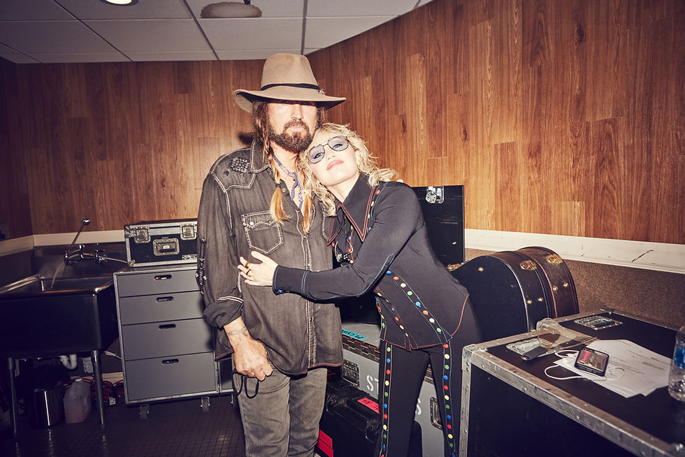 Miley Cyrus abraça Billy Ray Cyrus, ambos sorrindo, em bastidores com caixas de equipamento e paredes de madeira.