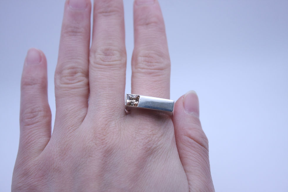 Thumbnail: Primer plano frontal de mano con anillo rectangular de plata y circonita naranja en caja negra de joyería. Try on.