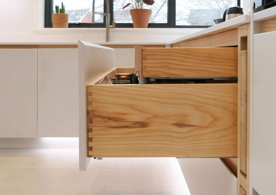 A handmade, modern kitchen with smooth white kitchen cabinetry, a bespoke waterfall worktop, ash wooden features, bespoke detailing and innovative storage solutions