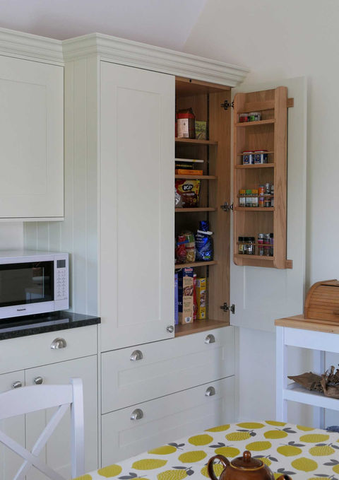 a handcrafted bespoke custom kitchen featuring shaker inspired doors and drawer fronts with a frameless modern design. The kitchen is painted in a soft, off-white colour and paired with stain nickel handles and a granite worktop. this design features innovative corner storage and slimline storage, a large oak larder cupboard and a classic Belfast sink with integrated kitchen appliances