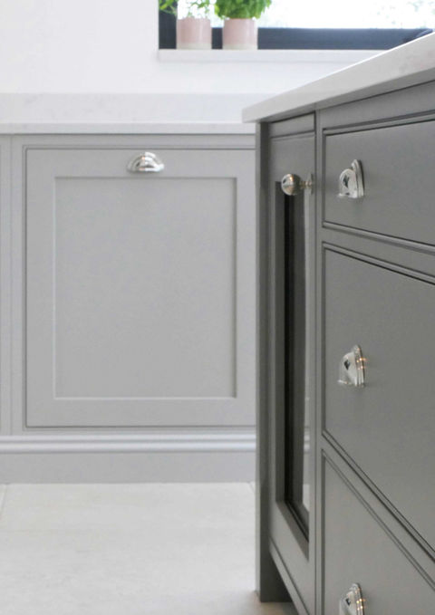 This handcrafted shaker kitchen features light grey shaker cabinetry around the edge of the space and a dark grey kitchen island in the centre of the design. The shaker-inspired cabinetry is paired with satin nickel handles and Carrara quartz worktops. This kitchen features plenty of you handmade oak details, a beautiful countertop cupboard, an integrated wine fridge with a glazed cabinetry door and handcrafted, bespoke cabinetry details.