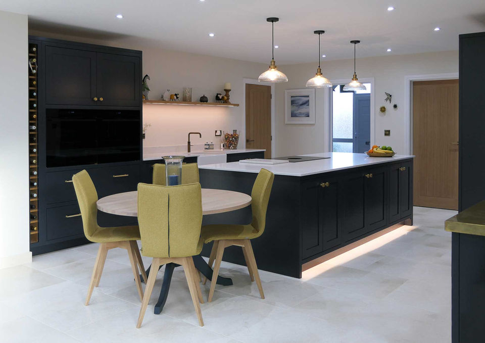 a handcrafted bespoke shaker kitchen painted in a dark grey colour with blue undertones and paired with satin brass kitchen handles and a quartz worktop. The large central island features several drawers and cupboards and unique plinth lighting. Several integrated appliances can be found around the kitchen along with a large kitchen larder and a freestanding bespoke dresser