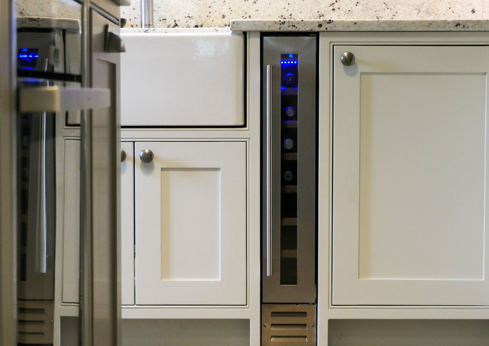 A handcrafted shaker-inspired kitchen with a small, social kitchen island. The kitchen cabinetry is painted in a soft white colour and paired with brushed nickel handles. This kitchen design features an integrated wine rack, two oak chopping boards, bespoke wall cabinetry and a beautiful countertop cupboard with oak shelving.