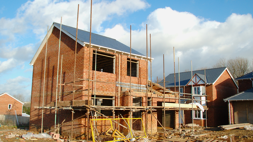 Brick house under construction with scaffolding, set in a suburban area with clear blue sky and clouds. Debris scattered around.