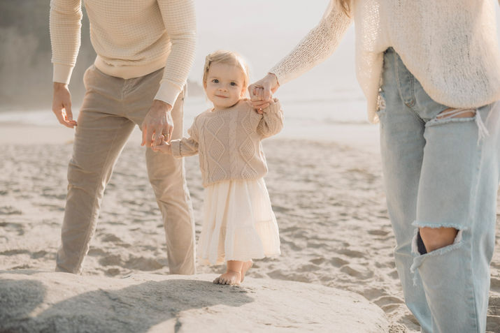 Toddler in beige cable-knit sweater balances on a coastal rock holding parents’ hands at golden hour on Dana Point beach.