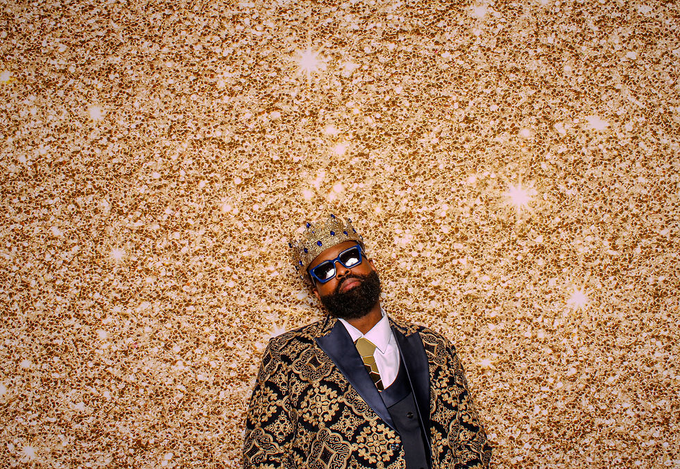 Black man wearing a gold crown, sunglasses, and a black-and-gold brocade suit, standing confidently against a glittering gold backdrop; conveys luxury, presence, and self-expression central to the Sole_Stys™ brand.