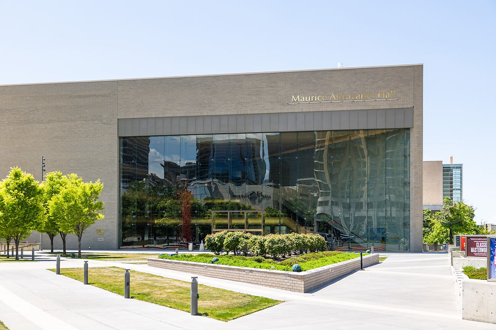 Modern building with large glass windows reflecting the city. Visible text: Maurice Abravanel Hall. Lush green trees and plants surround.
