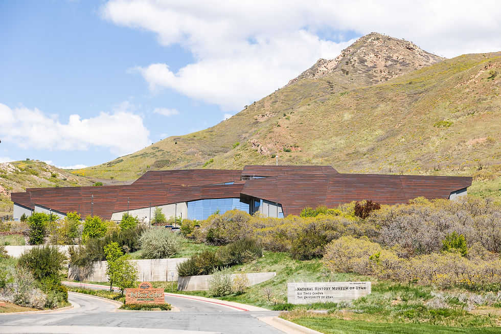 Modern building with angular brown roof set against a mountain backdrop. Greenery surrounds it. Sign reads Natural History Museum of Utah.